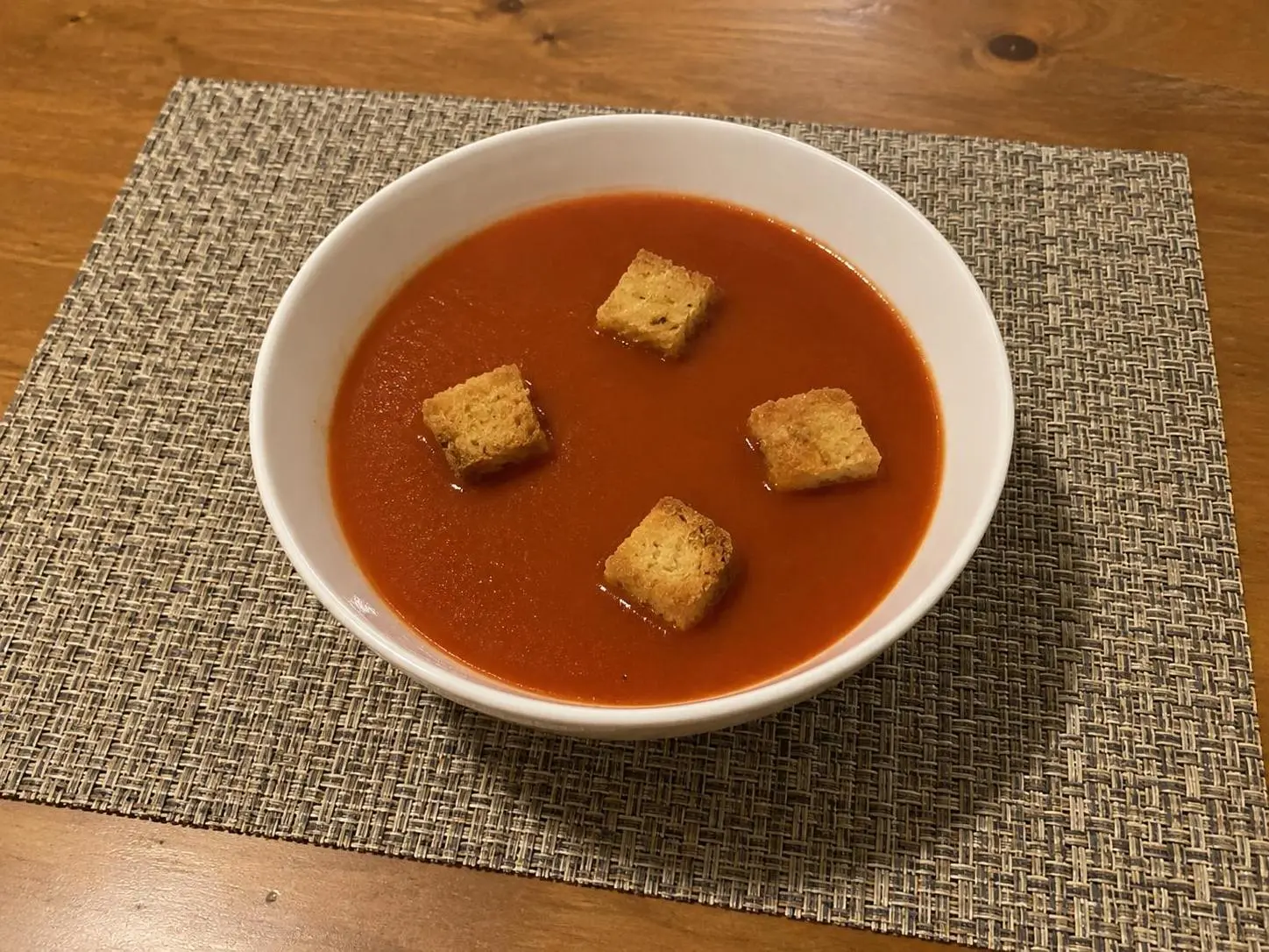 AI-made sample phone photo of tomato soup with four croutons on a kitchen table; generated demonstration, not a real customer dish.